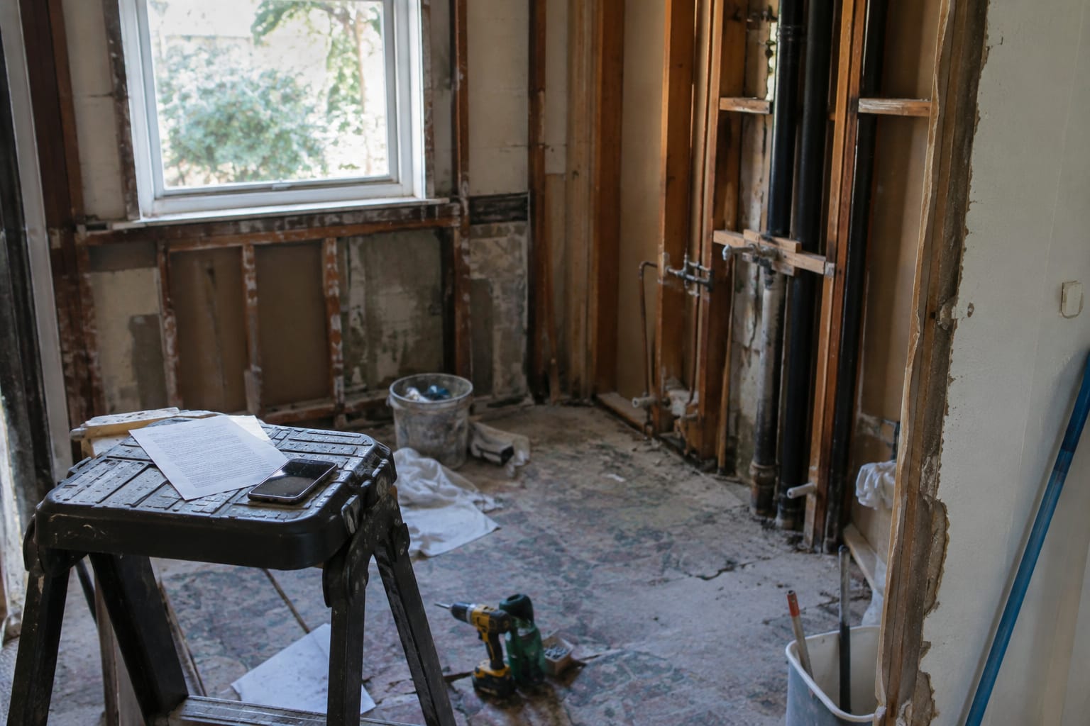 Salle de bain en chantier laissée à l'abandon, murs à nu et outils au sol, avec un devis et un téléphone posés sur un escabeau