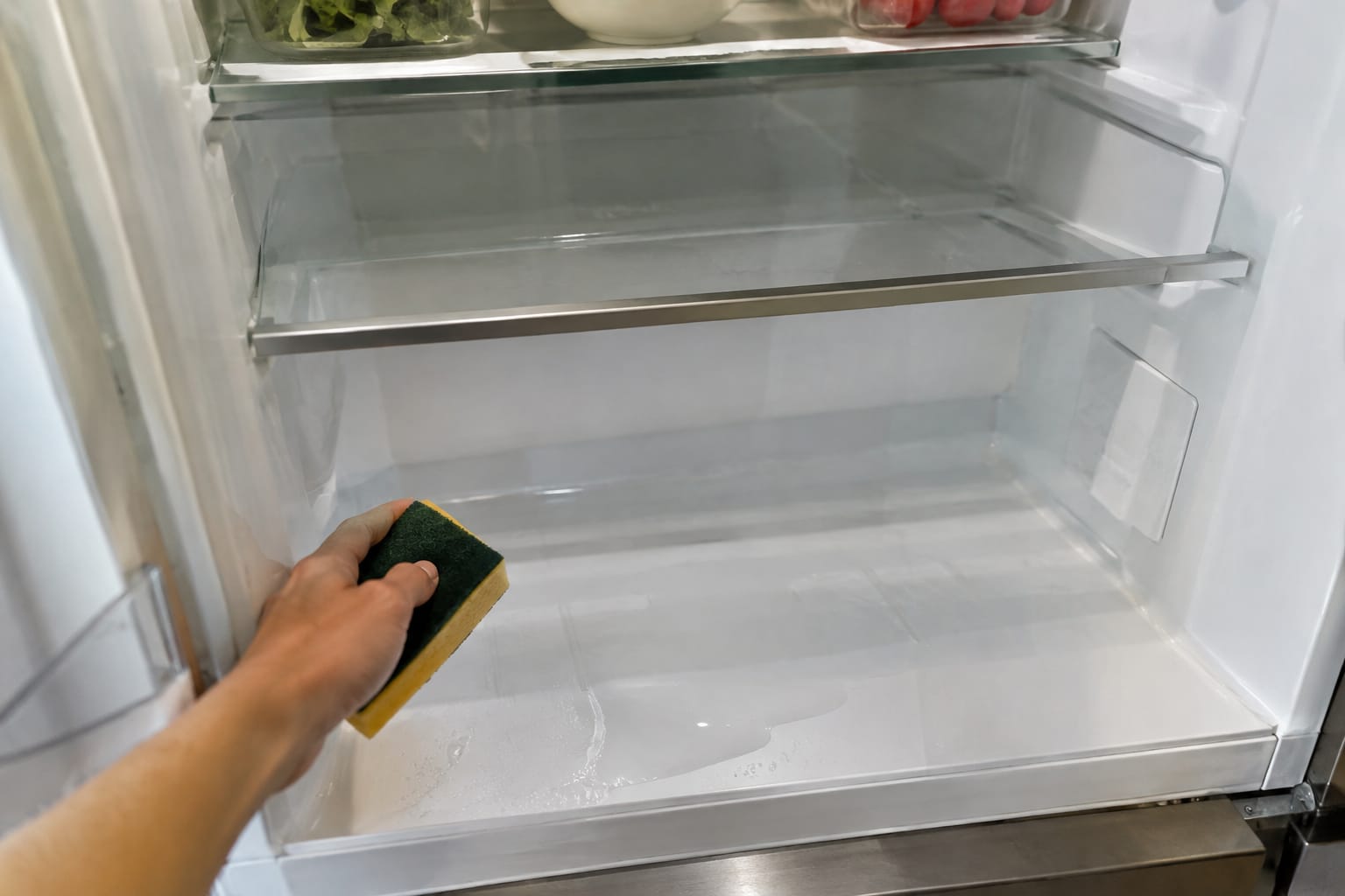 Main tenant une éponge à l'intérieur d'un réfrigérateur ouvert dont le bac à légumes a été retiré, une flaque d'eau au fond