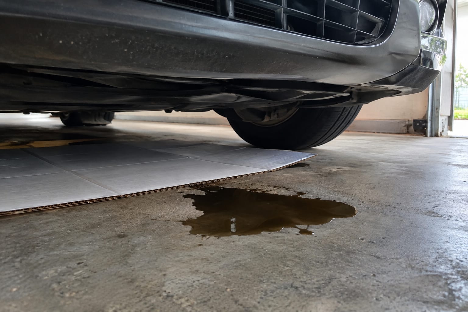 Petite flaque de liquide brun sur le sol en béton d'un garage sous l'avant d'une voiture, à côté d'un carton glissé sous le moteur